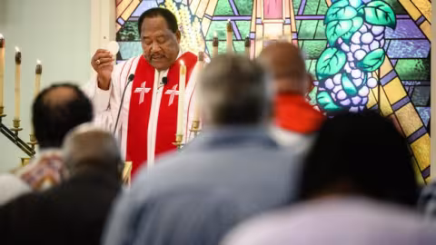 A clergyman in religious attire delivers a sermon in front of a stained glass window, while congregation members are seated, facing him attentively.