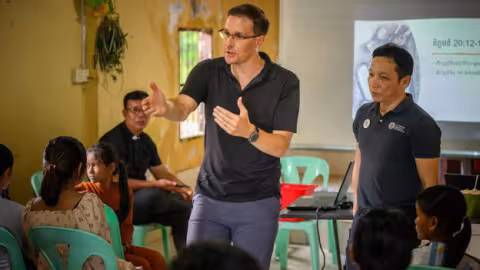 A group meeting in a community setting where a speaker gestures while explaining something to an audience. Others are seated, listening attentively. A presentation is visible in the background.